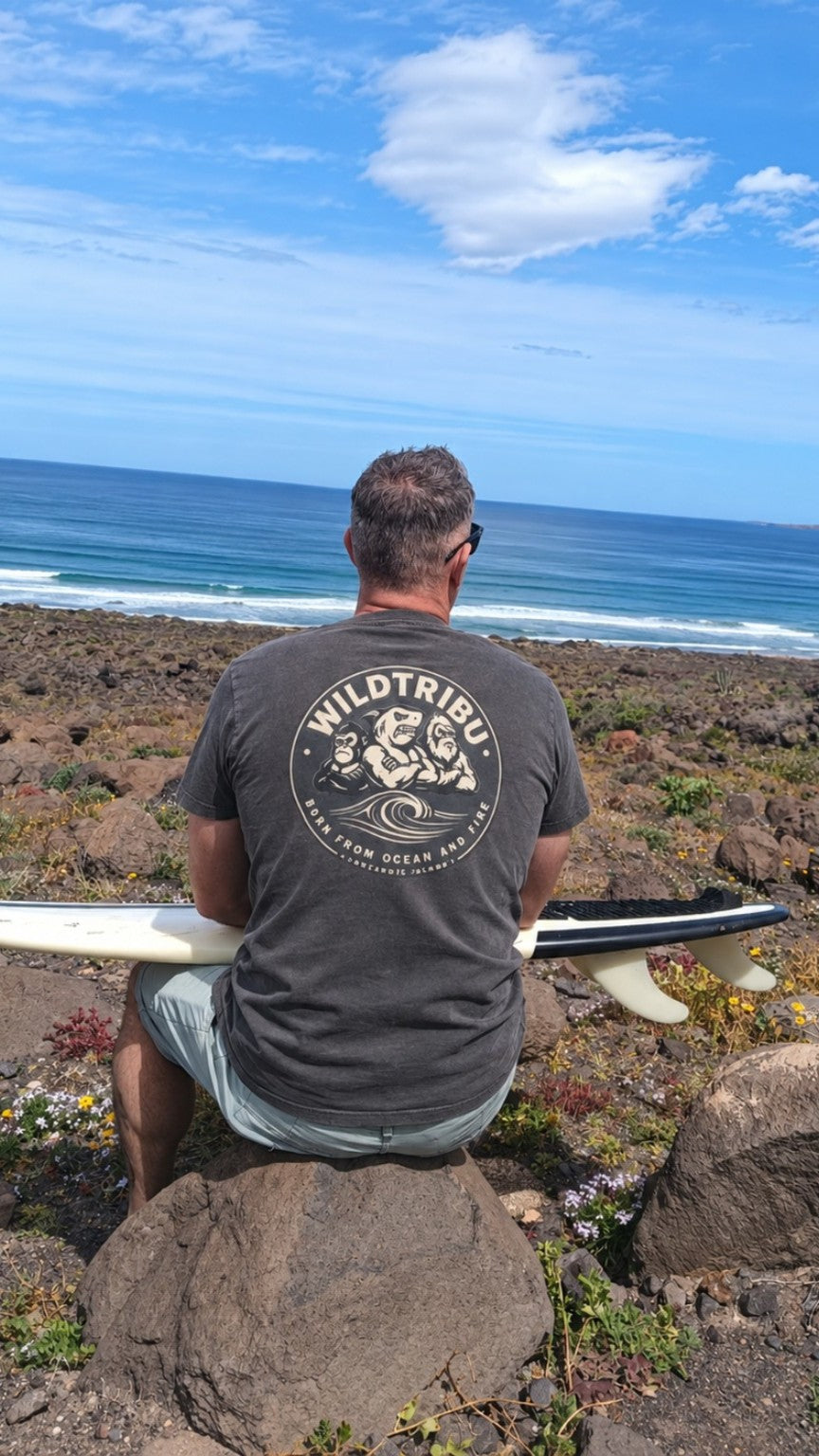 Camiseta Emblema de la Tribu en antracita, vista trasera con tabla de surf junto al océano