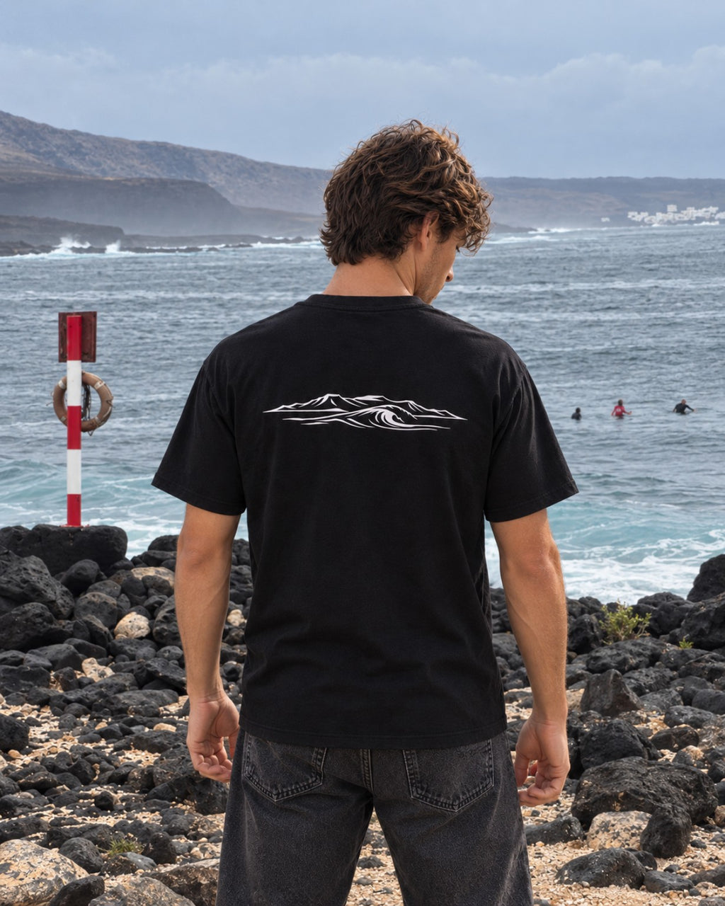 Camiseta Ocean and Fire negra con diseño de ola y volcanes en la espalda, junto al mar
